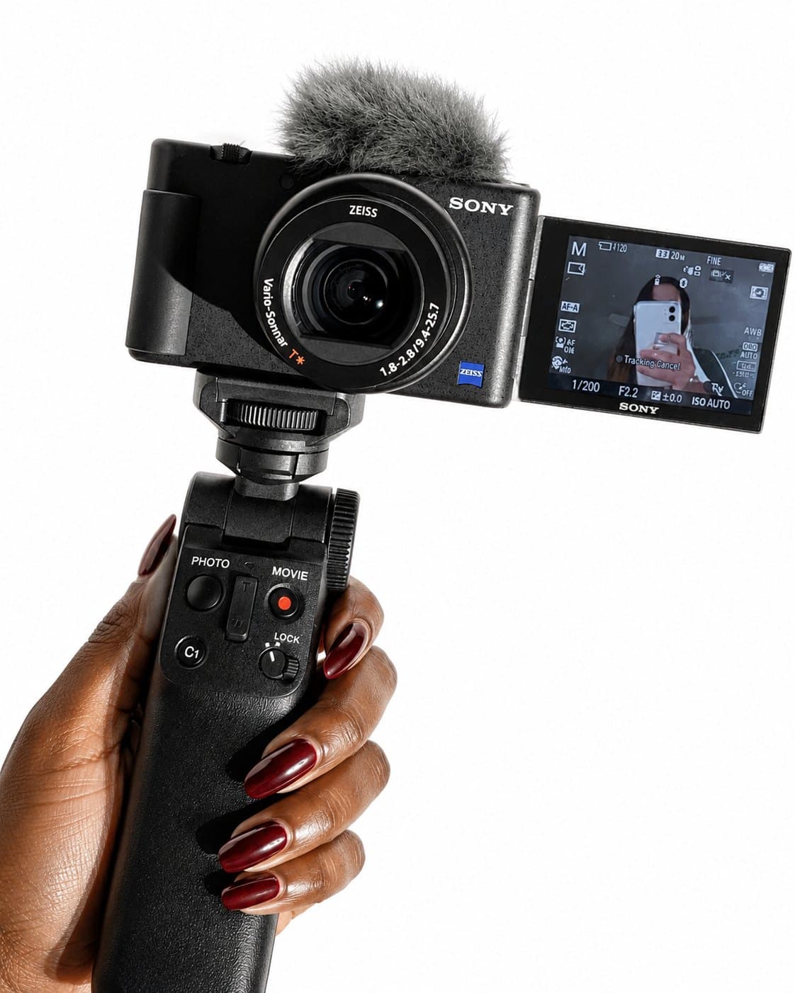 Hand with burgundy nails holding a compact vlogging camera on a grip, flip screen facing forward